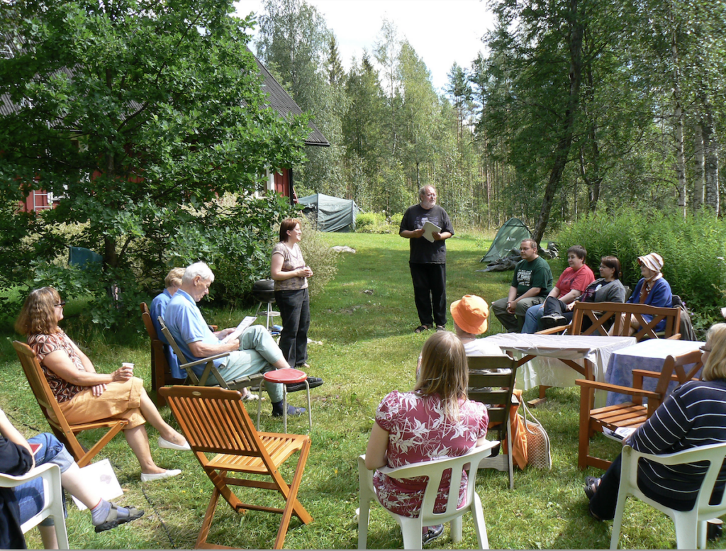 Usvan kesäleiri 2007: Anne Leinonen pitää puhetta vehreän pihapiirin keskellä, yleisö istuu ympärillä tuoleilla. Boris Hurtta seisoo taustalla paperit kädessään odottaen puheenvuoroa.